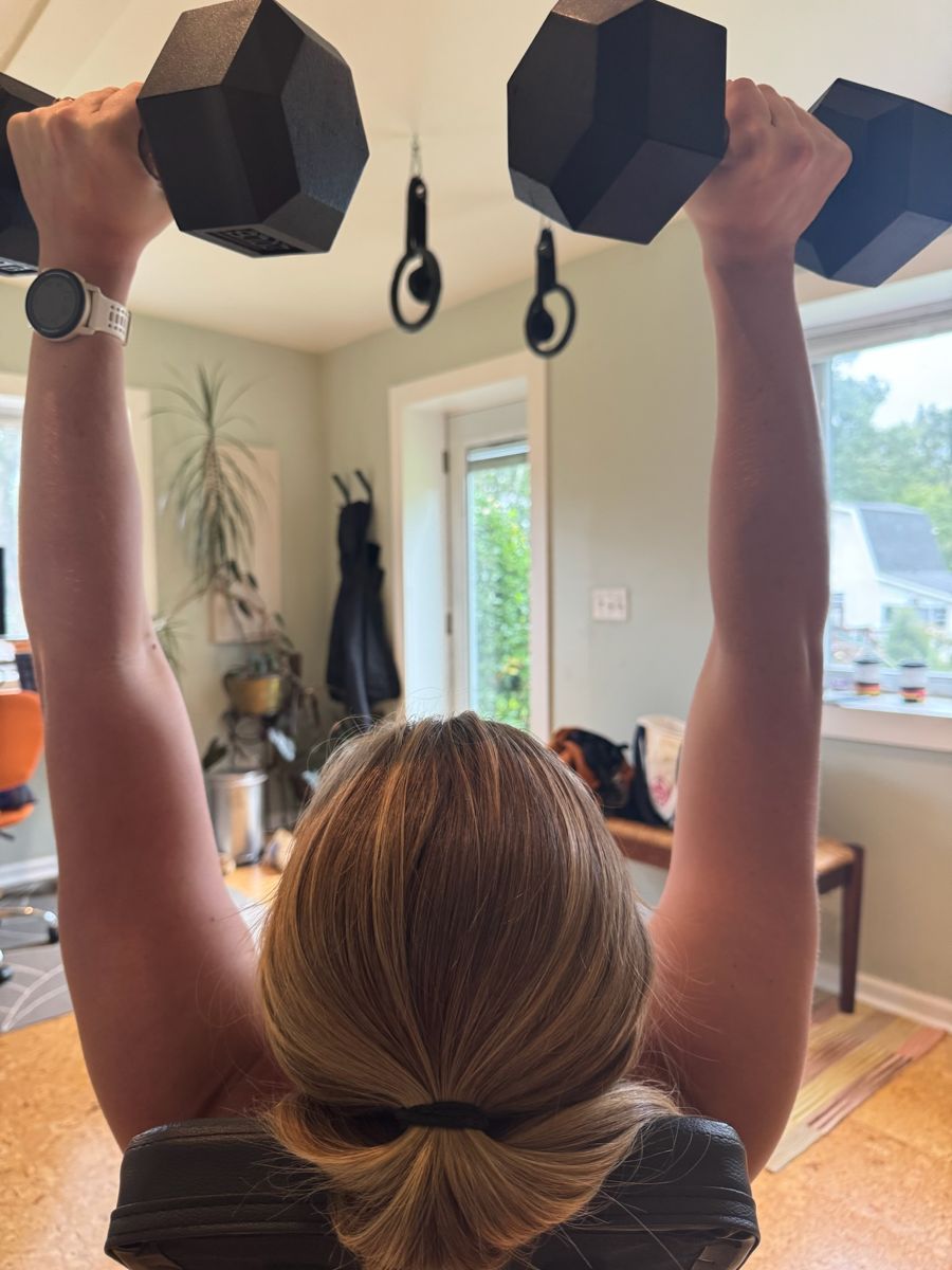 View from behind of Sadie pressing two dumbbells overhead, garden window beyond